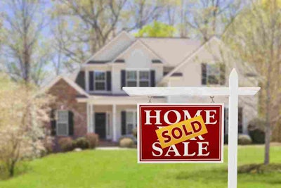 Photo of close up Home for Sale Sign with a yellow sold sticker and blurred house in the background with landscaped yard