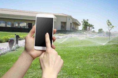 Photo of hands holding a smartphone while sprinkler system is watering green lawn in back ground