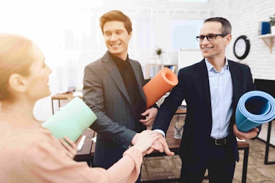 three employees in business attire holding yoga mats in one hand and the other hands reached out to each other in the center as to make a pact