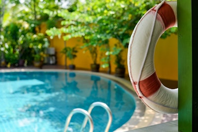 Close Up Of A White And Red Life Preserver Hanging On Green Wall With A Clear Blue Inground Swimming Pool In The Back Ground That Is Aligned With Potted Trees
