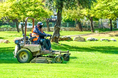 A landscaper wearing a black face mask while cutting grass on a commercial lawn mower