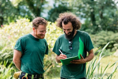 two landscapers wearing green shirts reviewing notes on a clipboard