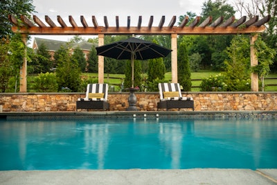 stone wall below a pergola and behind lounge chairs next to a pool