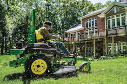Man operating green and yellow John Deere lawn mower on residential property with brick house and trees